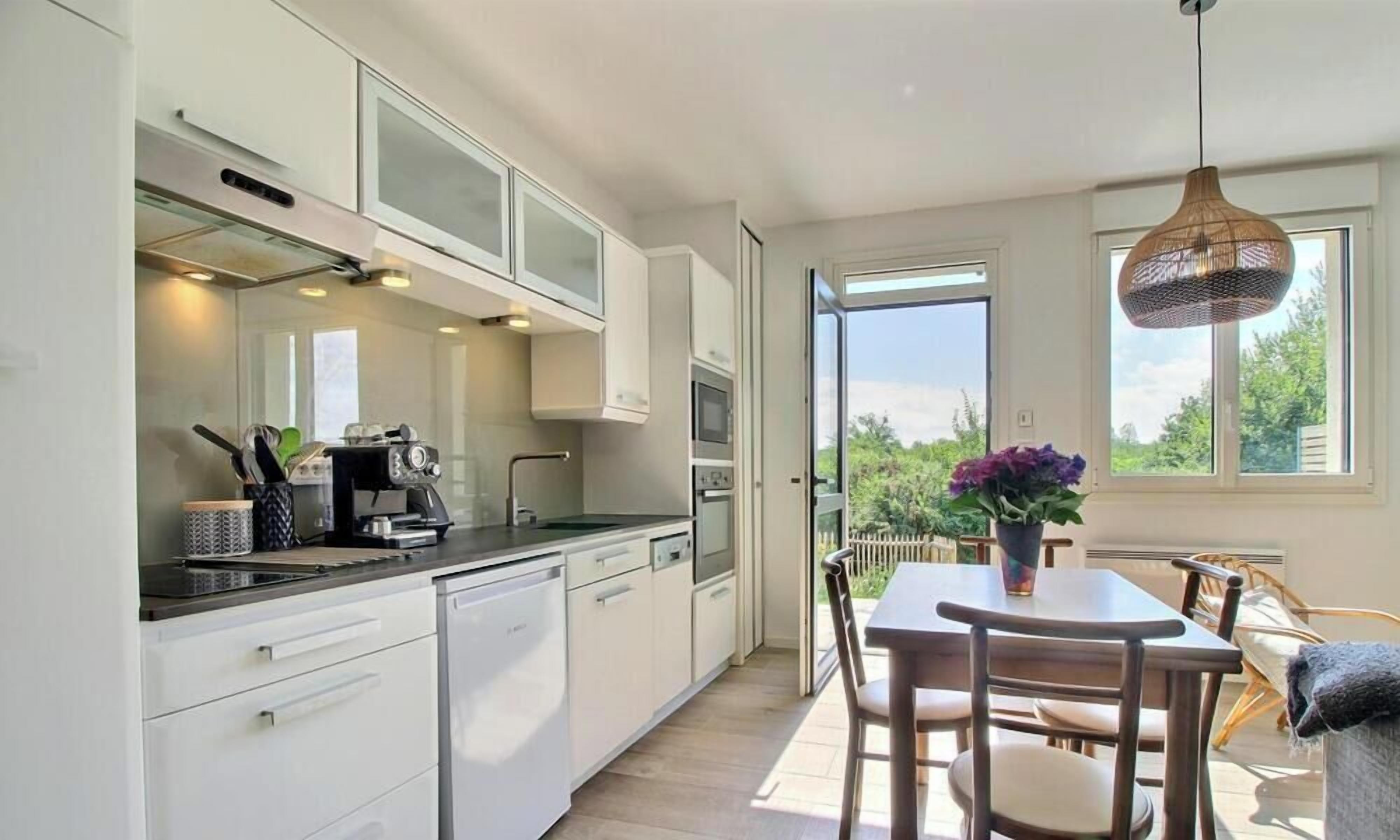 A modern kitchen with white cabinets, a built-in oven, and a coffee machine. There is a dining area with a table and chairs, and a large window providing a view of the outdoors.