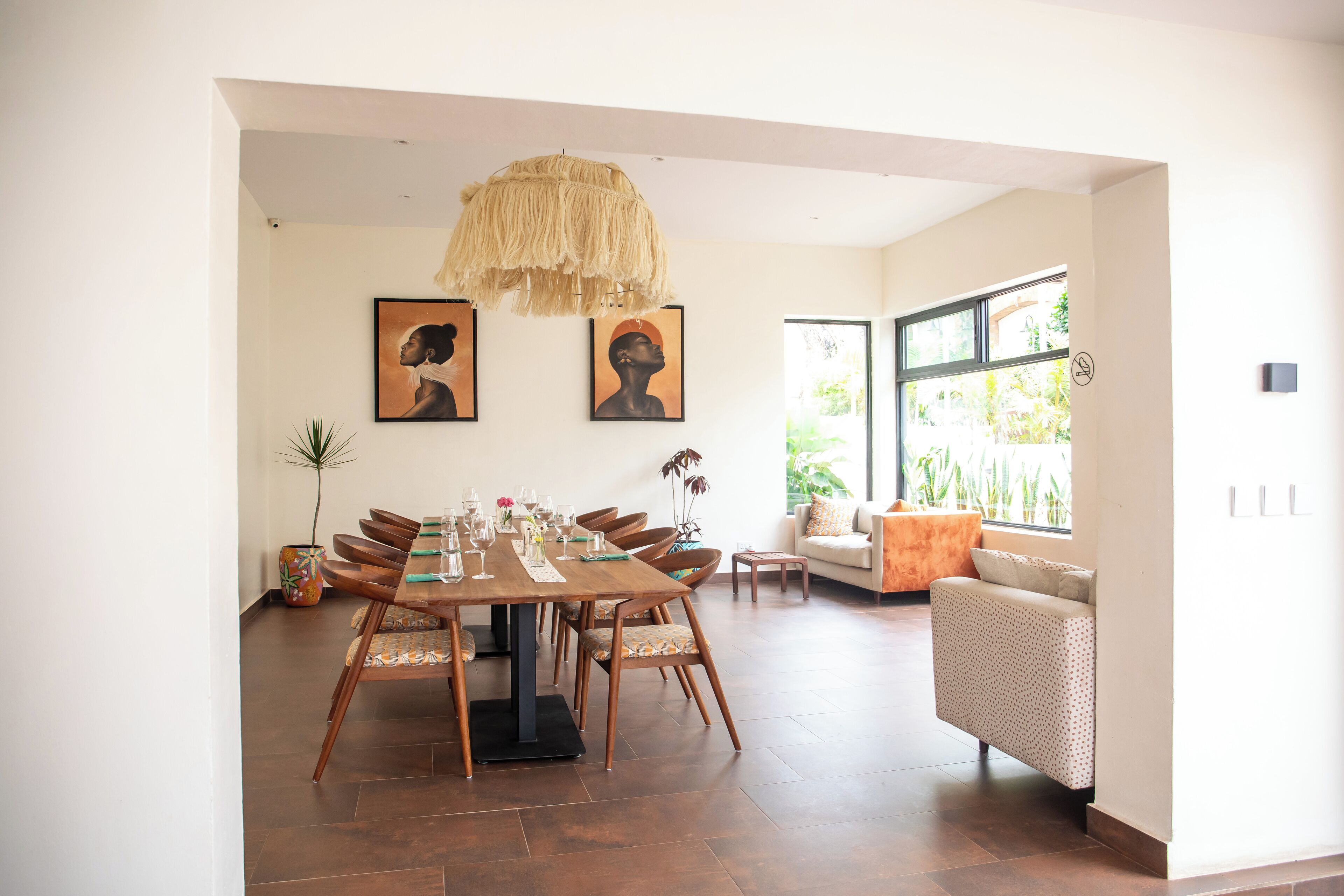A dining area with a wooden table set for four, surrounded by chairs. There are two framed artworks on the wall, a hanging light fixture, and a window with a view of greenery.