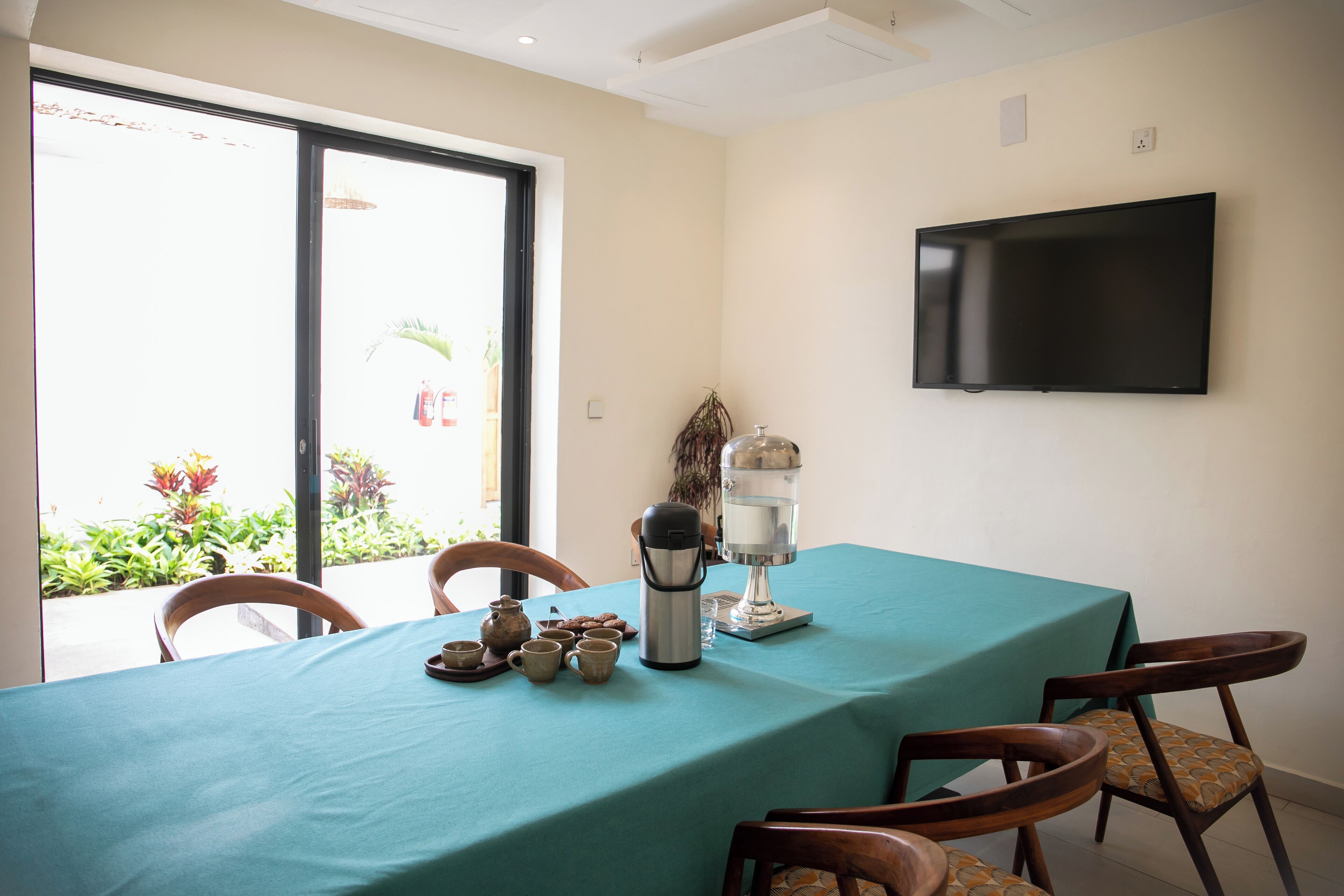 A dining area with a table covered by a teal tablecloth, wooden chairs, a coffee maker, and a water dispenser. A flat-screen TV is mounted on the wall.