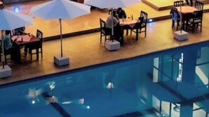 Un área junto a la piscina con mesas y sillas bajo sombrillas, una piscina con agua cristalina y una terraza de madera.
