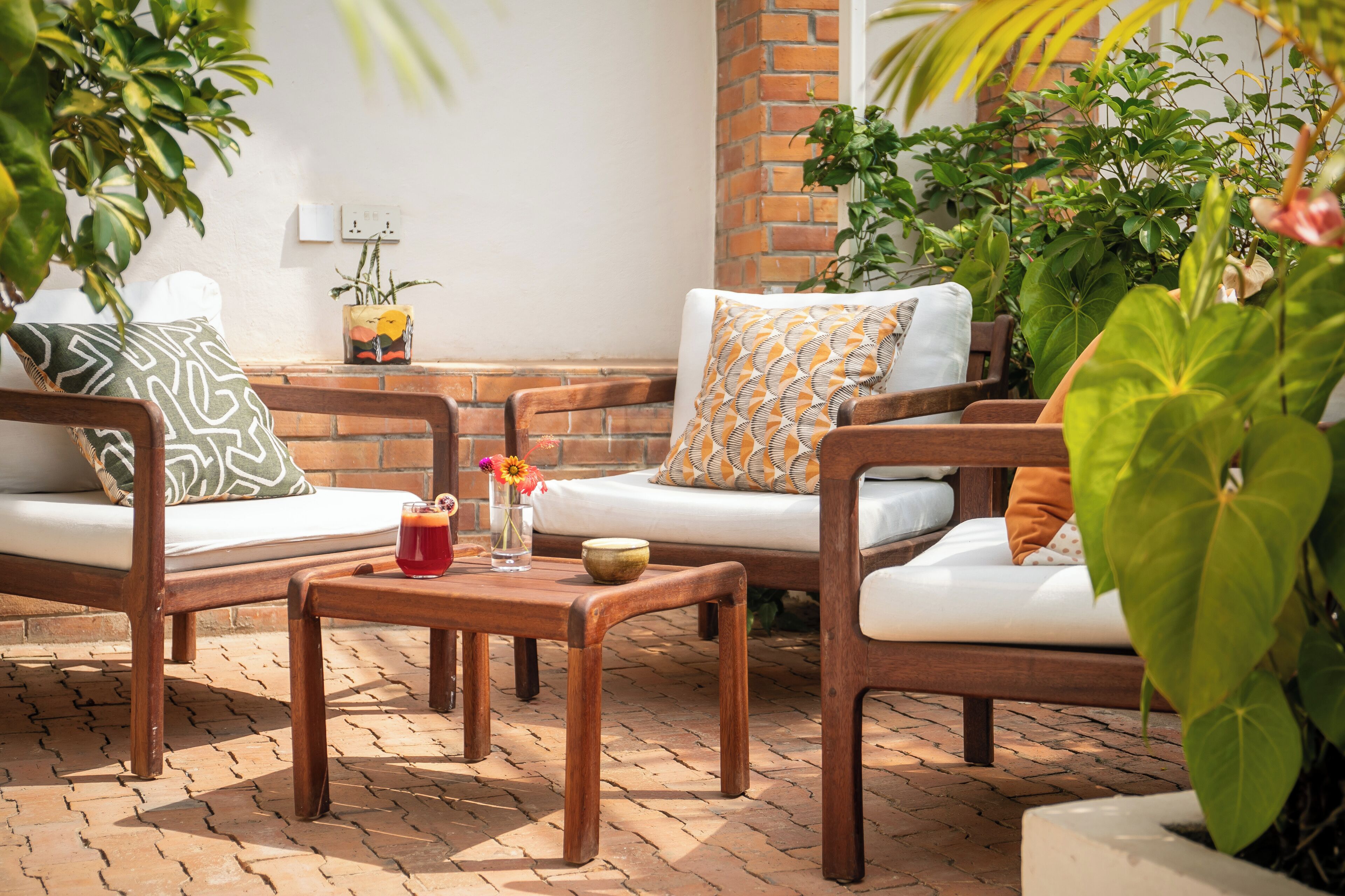 Un espace patio aménagé avec des meubles en bois, notamment un canapé et des fauteuils, une petite table avec une boisson et un vase de fleurs, ainsi que des plantes en pot.