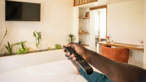 Una persona sosteniendo un control remoto en una habitación con una cama, un televisor montado en la pared y un escritorio con una silla.