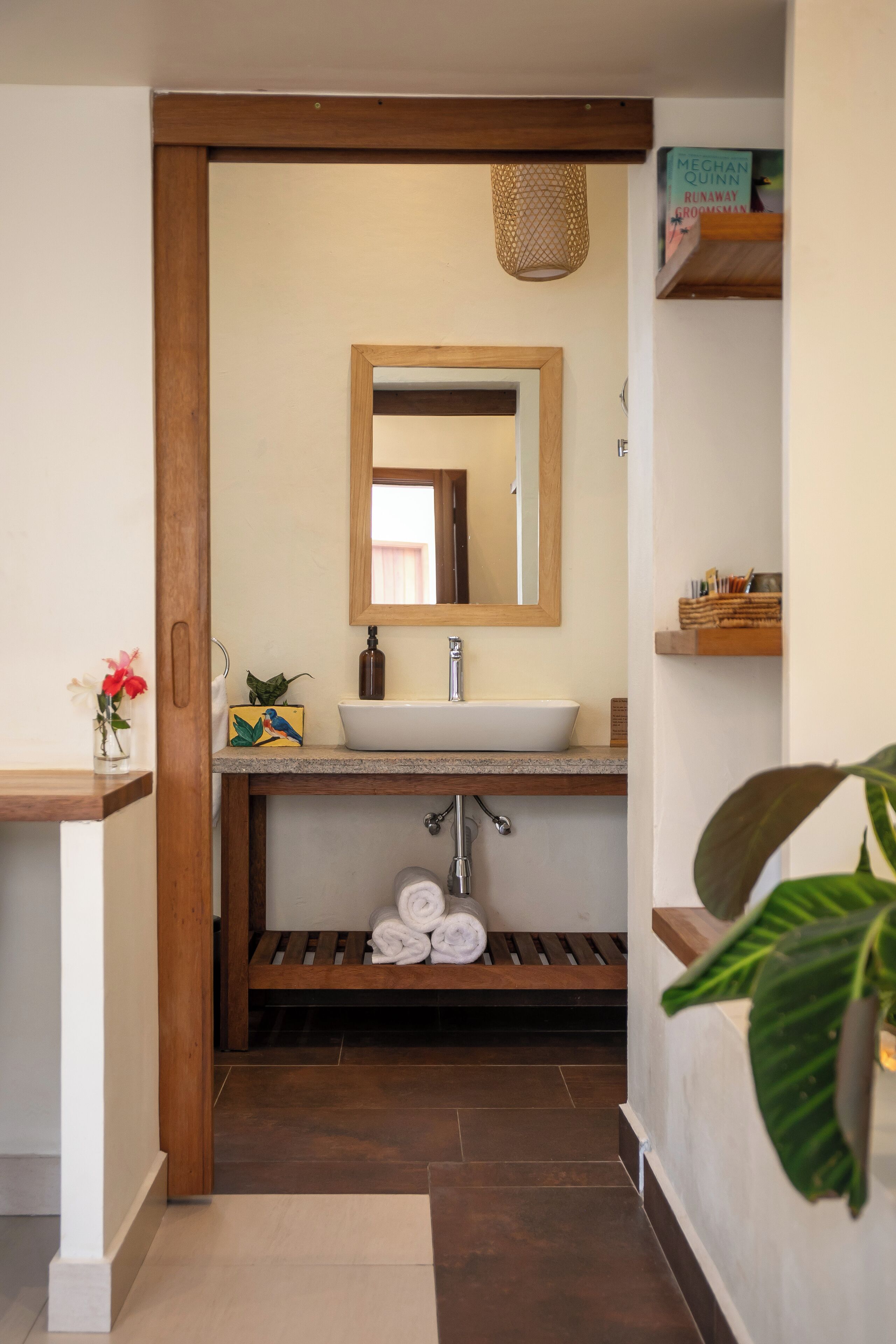 A bathroom with a wooden vanity, a white sink, a mirror, and a shelf with toiletries.