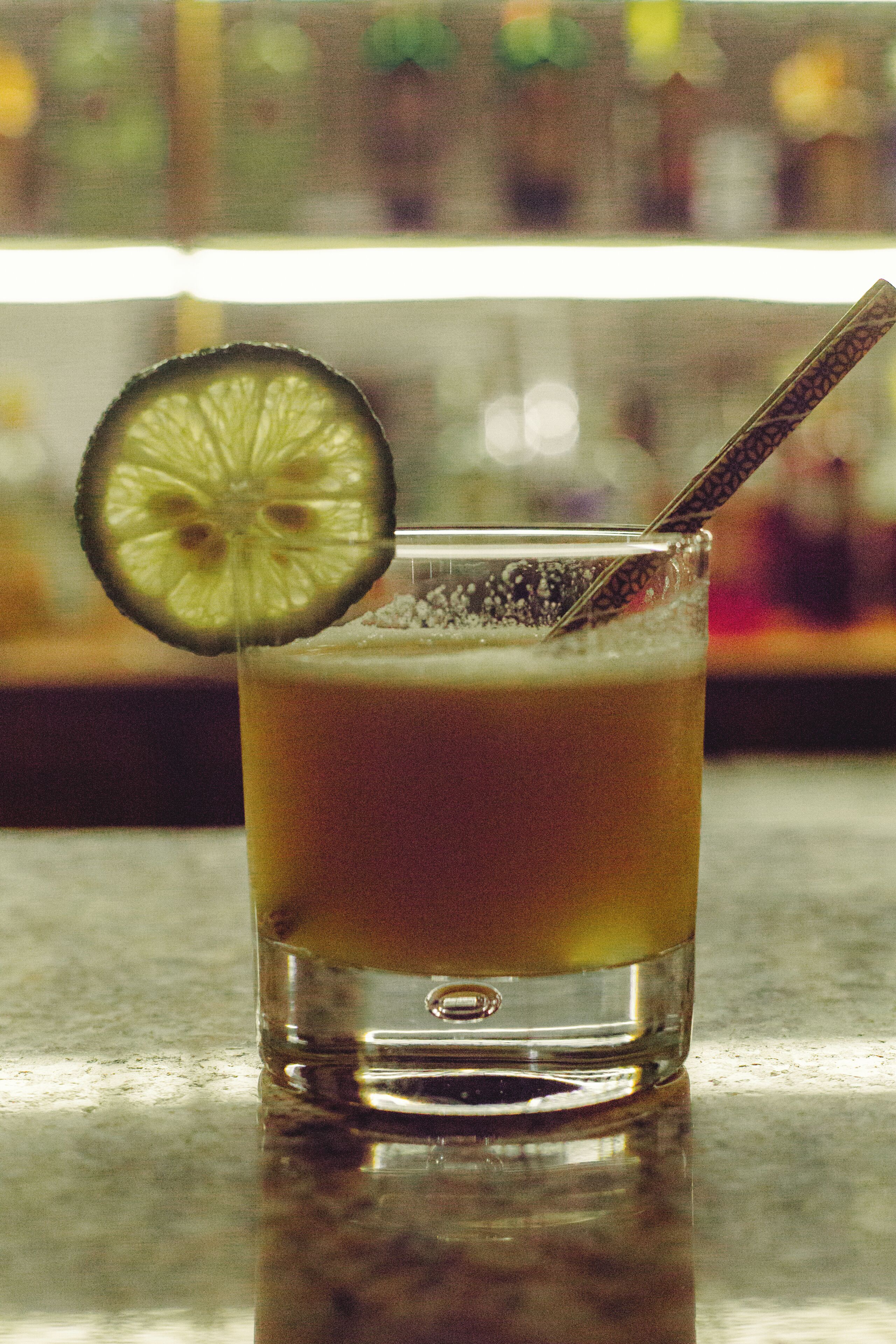 A glass of amber-colored beverage with a slice of lime and a straw, placed on a reflective surface.