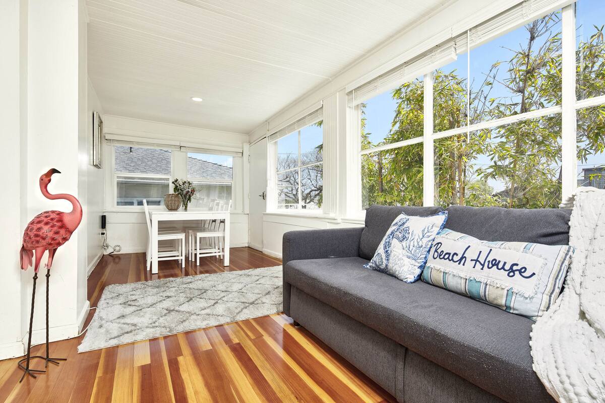A living room with a grey sofa, a 'Beach House' pillow, a red flamingo, and a dining area with white furniture.