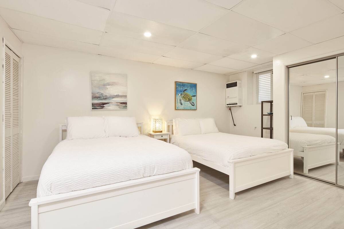 A bedroom with two white beds, a nightstand with a lamp, a painting on the wall, and a sliding glass door leading to another room.
