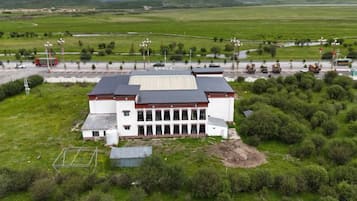 Um edifício grande com telhado plano e fachada simétrica, cercado por um campo gramado e uma estrada com veículos.