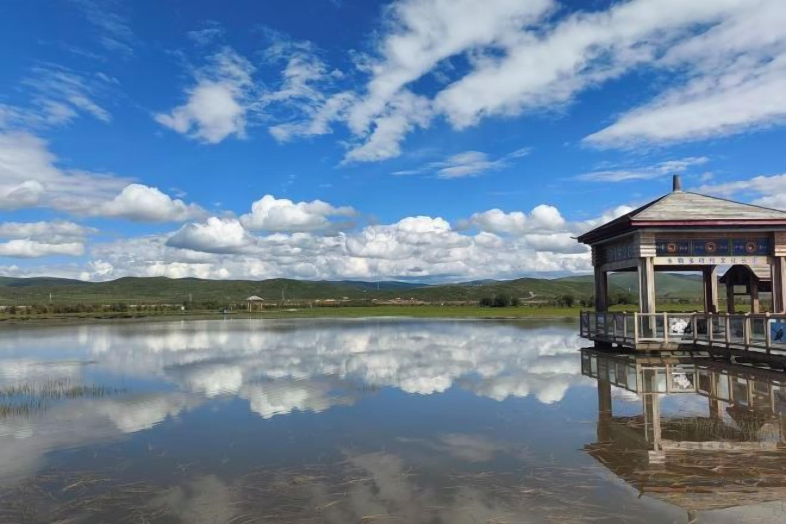 Un reflet net d’un bâtiment sur une étendue d’eau calme, avec un ciel dégagé et des collines lointaines.