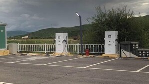 A parking area with two electric vehicle charging stations, a fence, and a green building in the background.