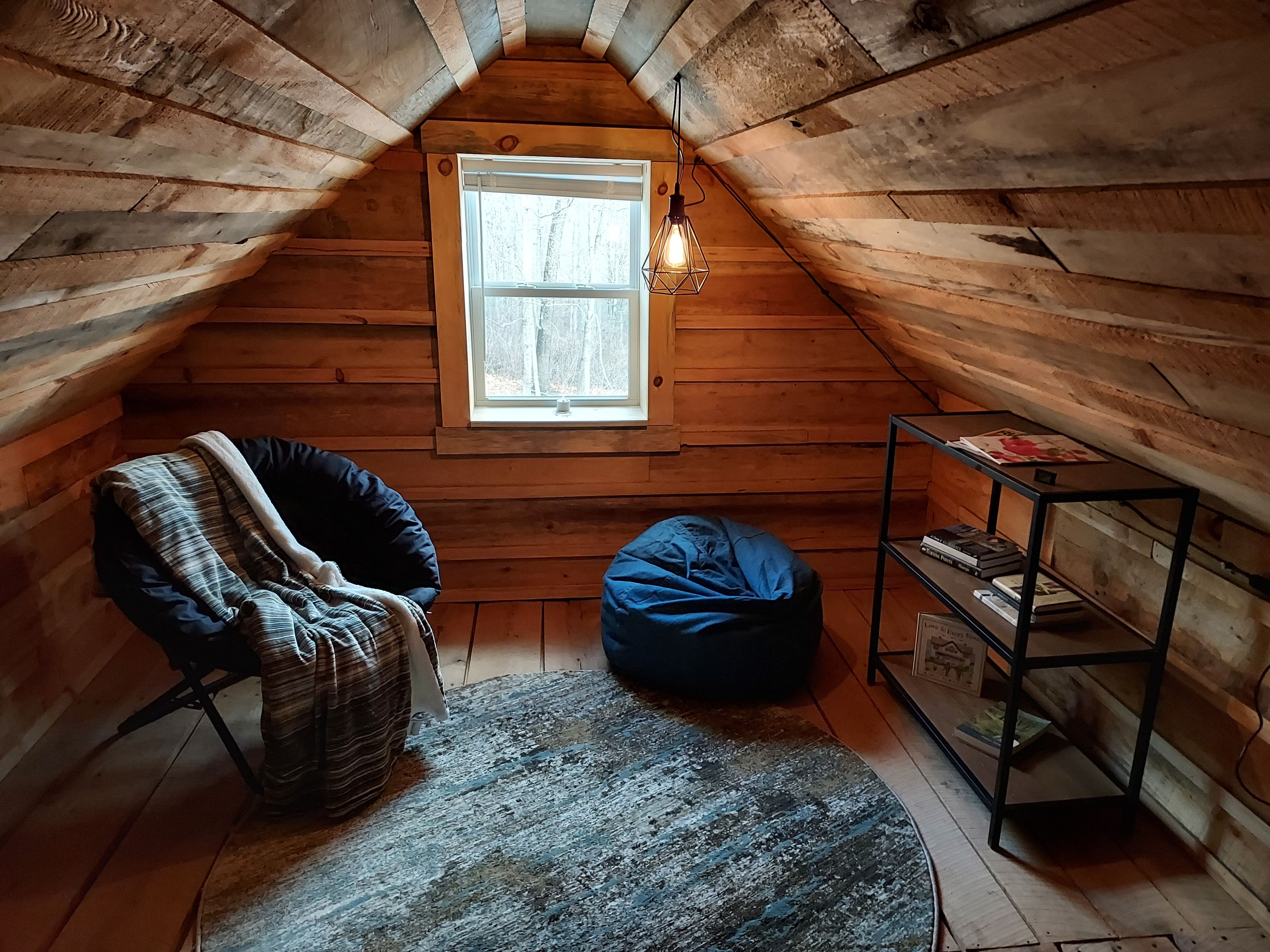 Una stanza accogliente in mansarda con pareti in legno, una finestra, un pouf, una sedia e una mensola metallica con dei libri.