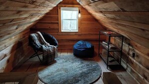 A cozy attic room with wooden walls, a window, a round rug, a bean bag chair, and a small table.