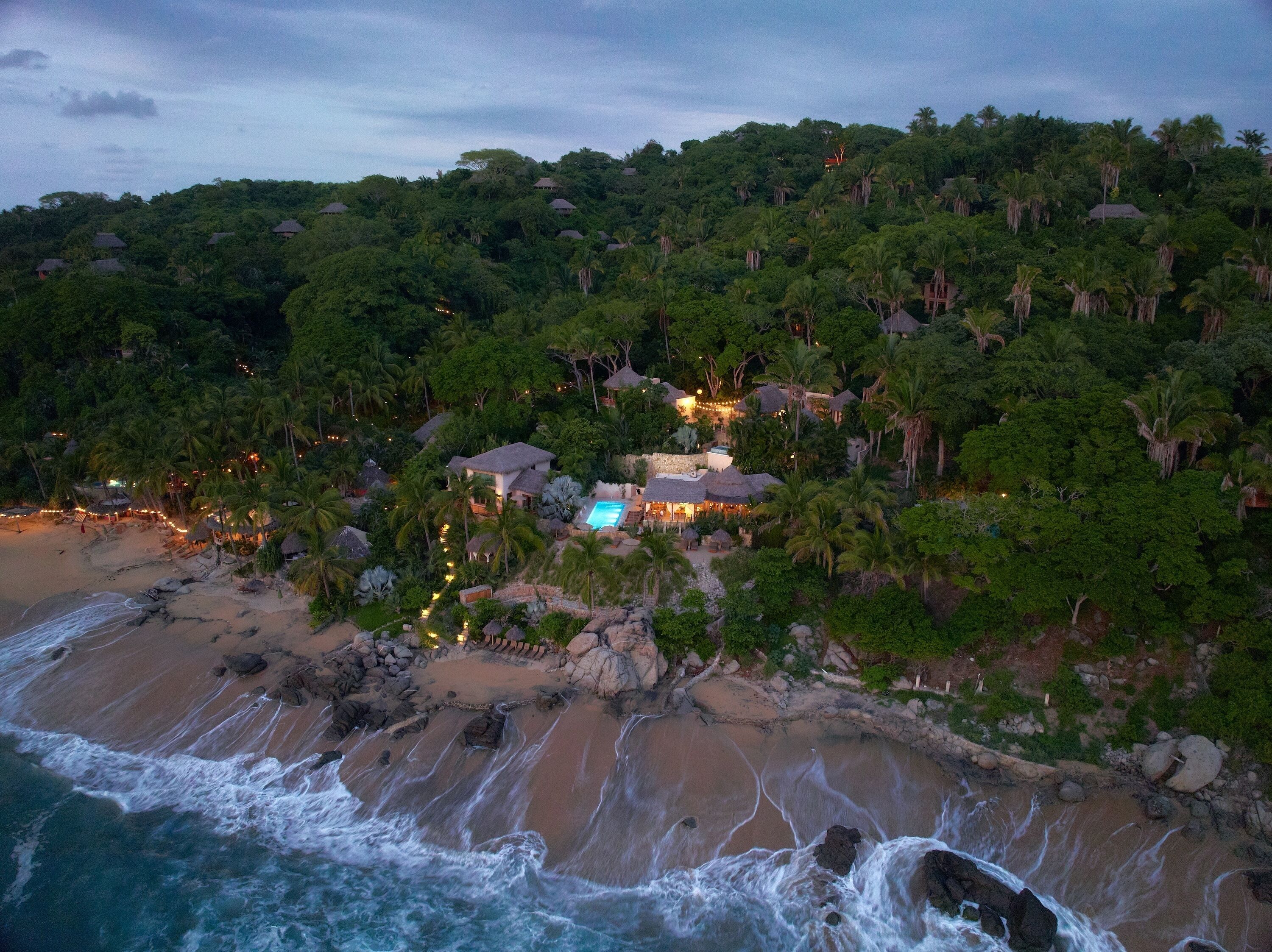 Un resort costero con varias edificaciones de techo de paja, una piscina y una playa.