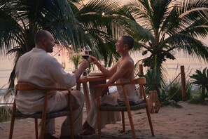 Dos personas sentadas en una mesa de madera con una copa de vino, bajo palmeras en una playa arenosa.