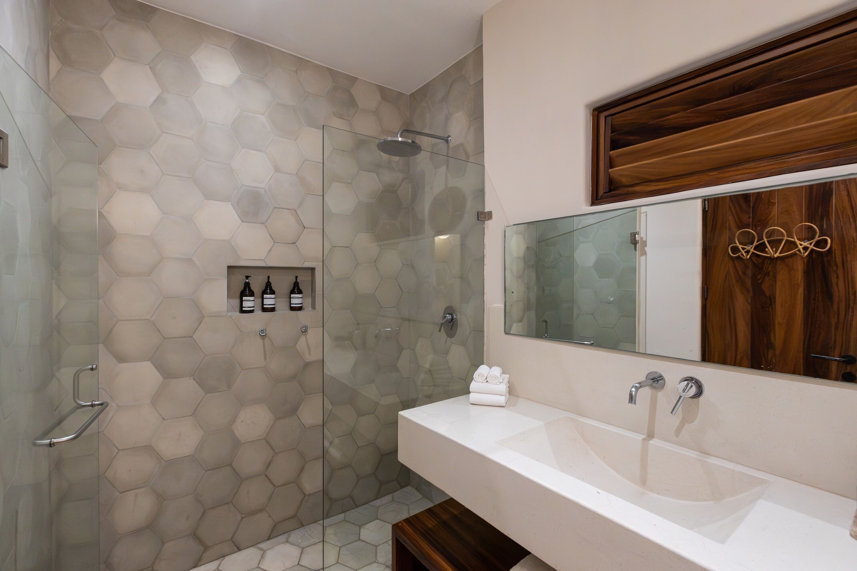Un baño con pared de azulejos hexagonales, cabina de ducha de vidrio, tocador de madera con espejo y barra para toallas.