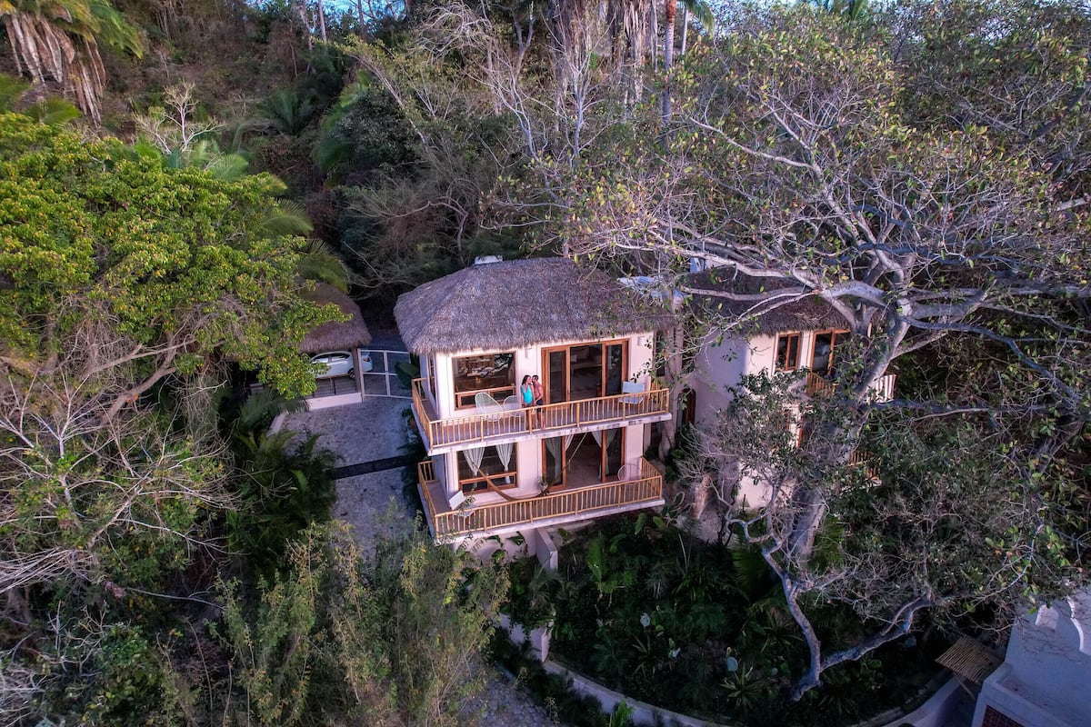 A two-story building with a thatched roof, surrounded by dense foliage.