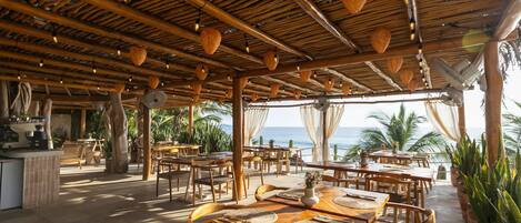 An outdoor dining area with wooden tables and chairs, a bar area, and a view of the ocean.