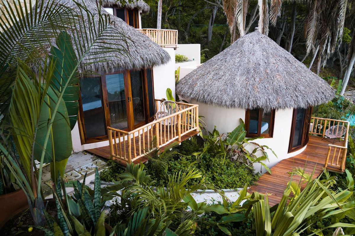 A thatched-roof bungalow with a wooden deck, surrounded by lush tropical vegetation.