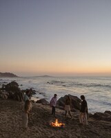 Un grupo de personas reunidas alrededor de una fogata en una playa rocosa, con el océano al fondo.