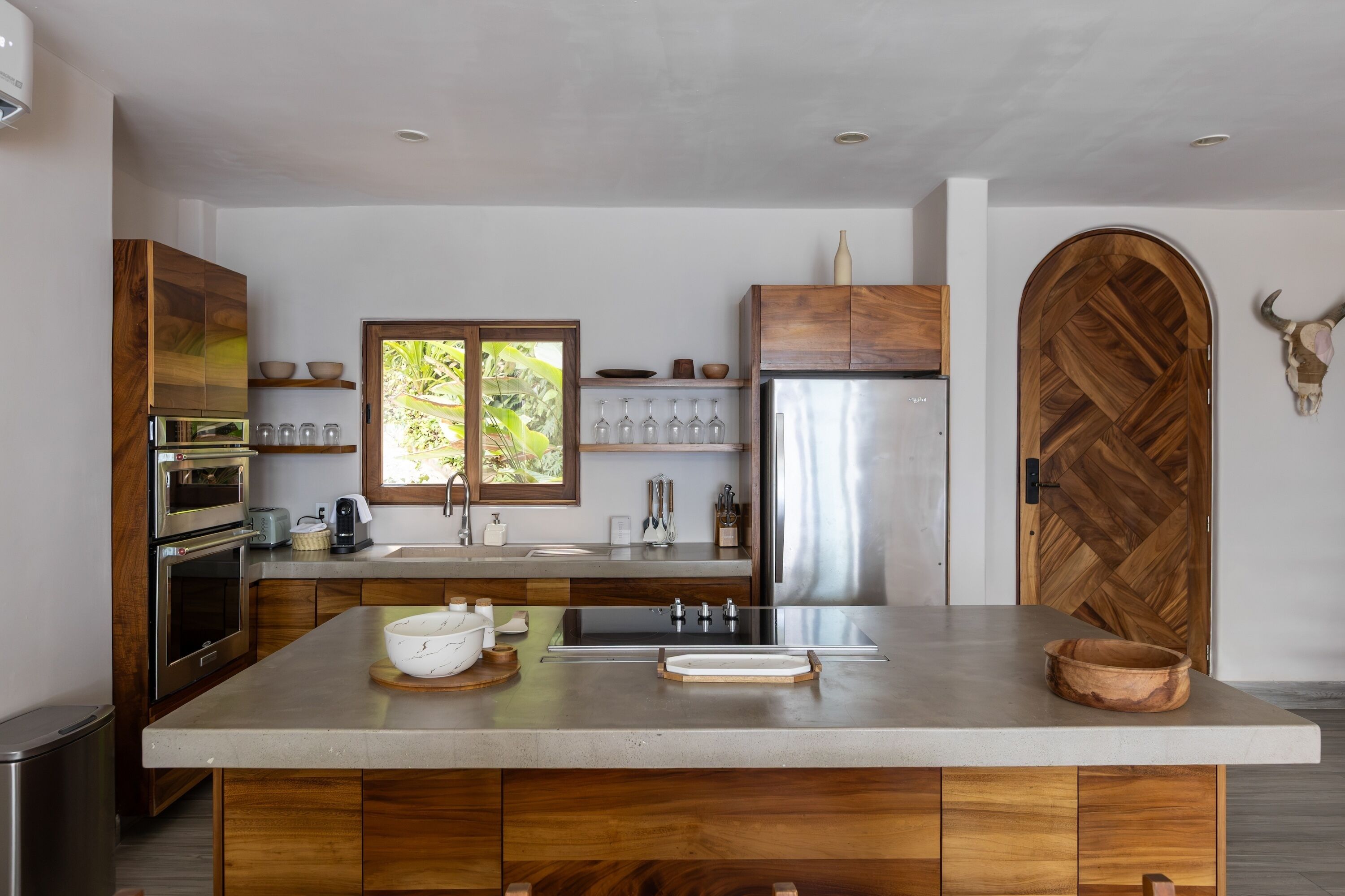 Une cuisine moderne dotée d’un grand îlot central, d’appareils électroménagers en acier inoxydable, de meubles de rangement en bois et d’une porte en bois originale ornée d’une décoration représentant un crâne de taureau.