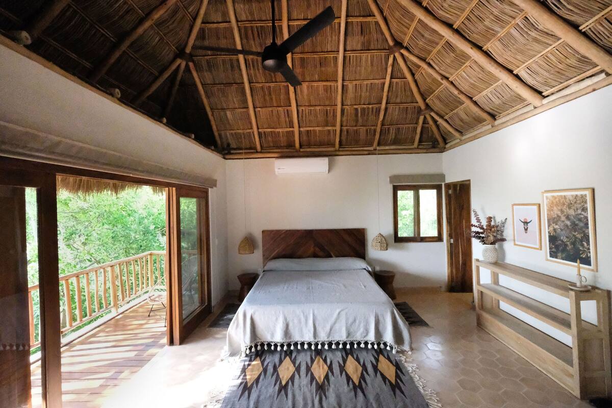 A bedroom with a bed, a ceiling fan, a wooden headboard, a bench, and a balcony with a railing.