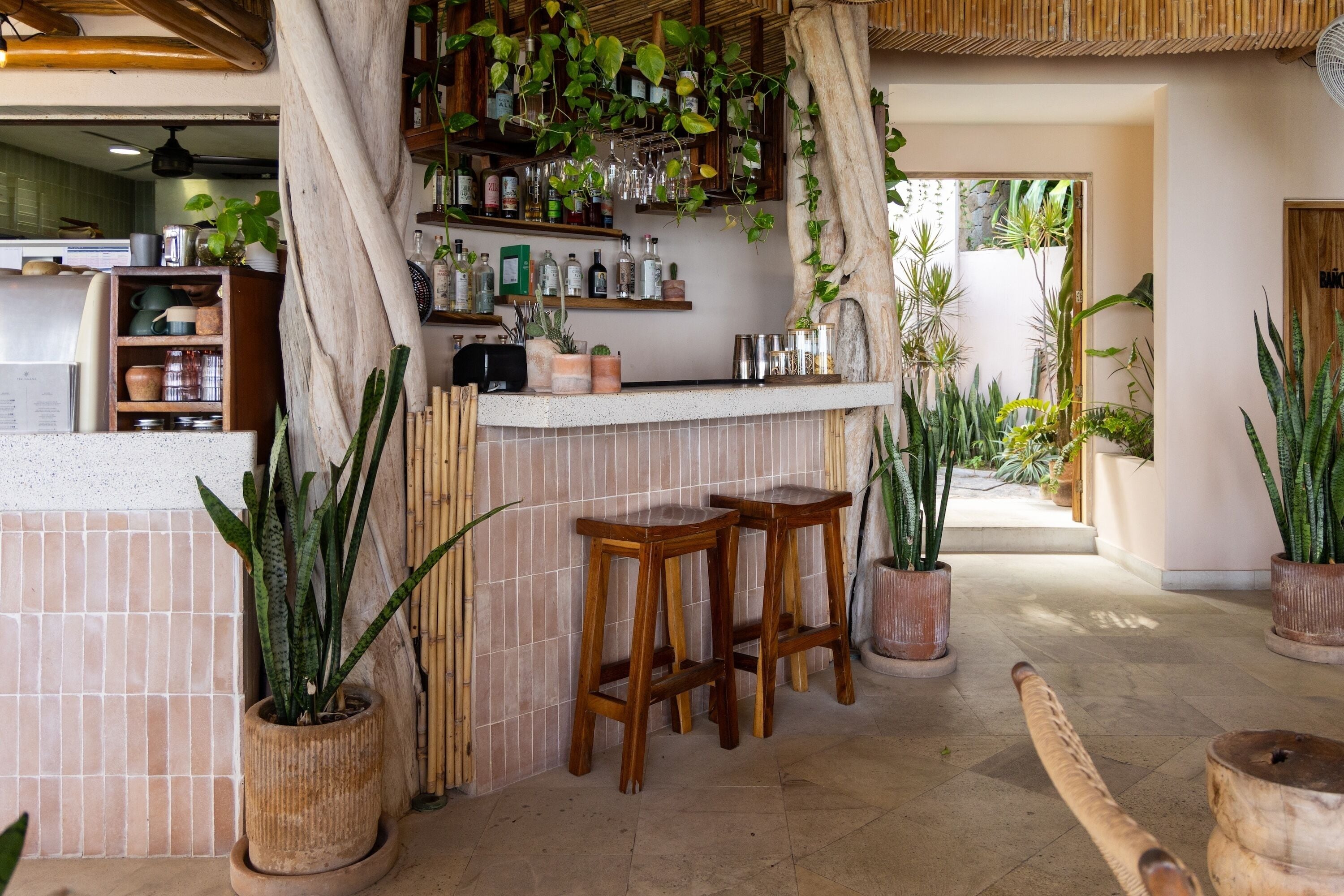 Uma área de bar com banquetas de madeira, painel de azulejos e diversas plantas.