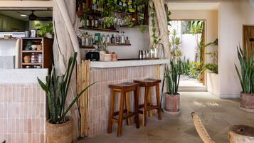 Uma área de bar com banquetas de madeira, painel de azulejos e diversas plantas.
