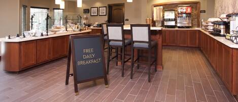 A hotel breakfast area with a sign stating 'FREE DAILY BREAKFAST,' a counter with a coffee machine, and seating with a view of the city.