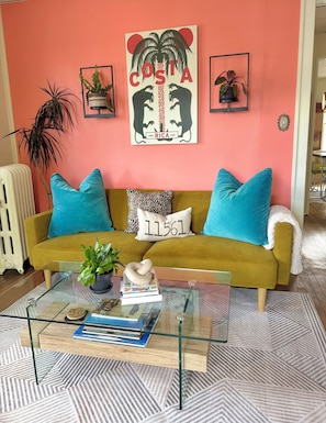 A living room with a mustard yellow sofa, teal cushions, a glass coffee table with books and a plant, a framed 'COSTA' artwork, and a radiator. - Private 2nd story apartment in the heart of Long Beach- Pura Vida! (Long Beach)