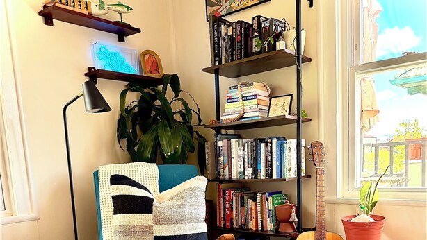 A cozy room with a blue armchair, a bookshelf, a guitar, and a window with a view. - Private 2nd story apartment in the heart of Long Beach- Pura Vida! (Long Beach)