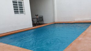 A rectangular swimming pool with a tiled edge, a potted plant, and a table with chairs in the background.