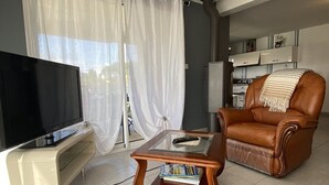 Een woonkamer met een flatscreen televisie, een salontafel met glazen blad en tijdschriften, een leren fauteuil, en een keuken in de achtergrond.