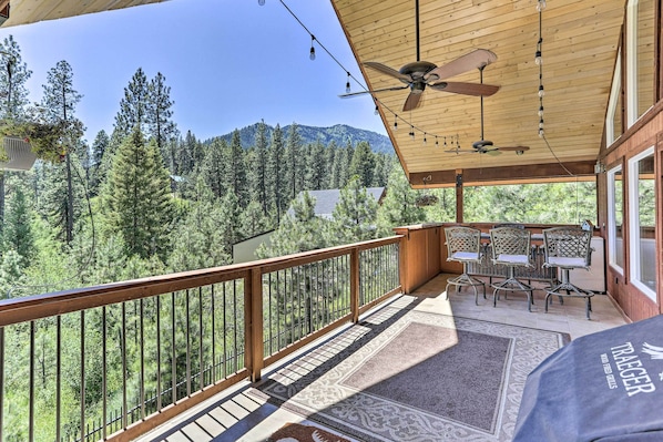 Terraza de madera con vistas a un bosque y montañas, ventilador de techo y mobiliario exterior.