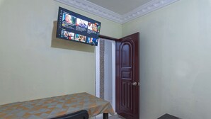 A small room with a bed, a TV mounted on the wall, a wooden door, and a small table with a remote control. - Sunset Single Room Near Airport Wi-fi & Parking (Santo Domingo Este)