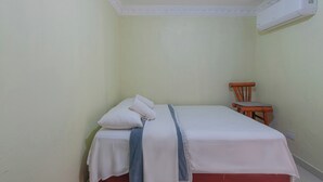 A small, clean room with a single bed, a wooden chair, and a wall-mounted air conditioning unit. - Sunset Single Room Near Airport Wi-fi & Parking (Santo Domingo Este)