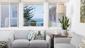 A living room with a grey sofa, a small side table, and a view of the ocean through large windows. - Moana Blue at Shoal Bay (Shoal Bay)