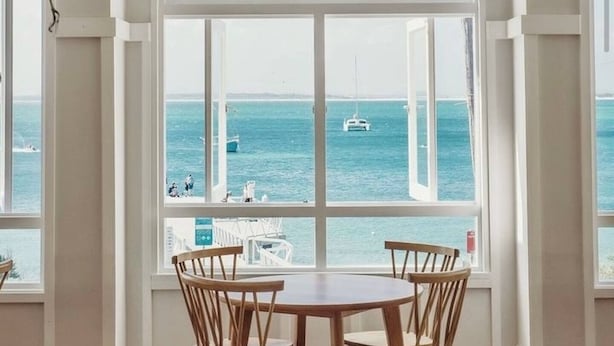 A room with a large arched window overlooking the sea, a ceiling fan, and a round table with chairs. - Moana Blue at Shoal Bay (Shoal Bay)