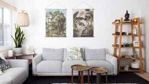 A living room with a grey sofa, two side tables, a wooden bookshelf, and tropical-themed wall art. - Moana Blue at Shoal Bay (Shoal Bay)
