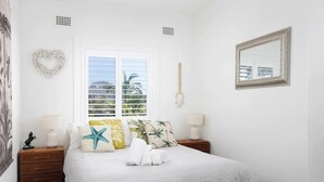 A bedroom with a bed, two bedside tables, a window with blinds, a heart-shaped decoration, and a mirror on the wall. - Moana Blue at Shoal Bay (Shoal Bay)