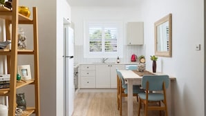 A modern kitchen with white cabinets, a dining area with a table and chairs, a window with blinds, and a wooden shelf with decorative items. - Moana Blue at Shoal Bay (Shoal Bay)