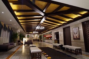 Un vestíbulo de hotel con techo alto, vigas de madera y un candelabro. Hay zonas de estar con mesas y sillas.