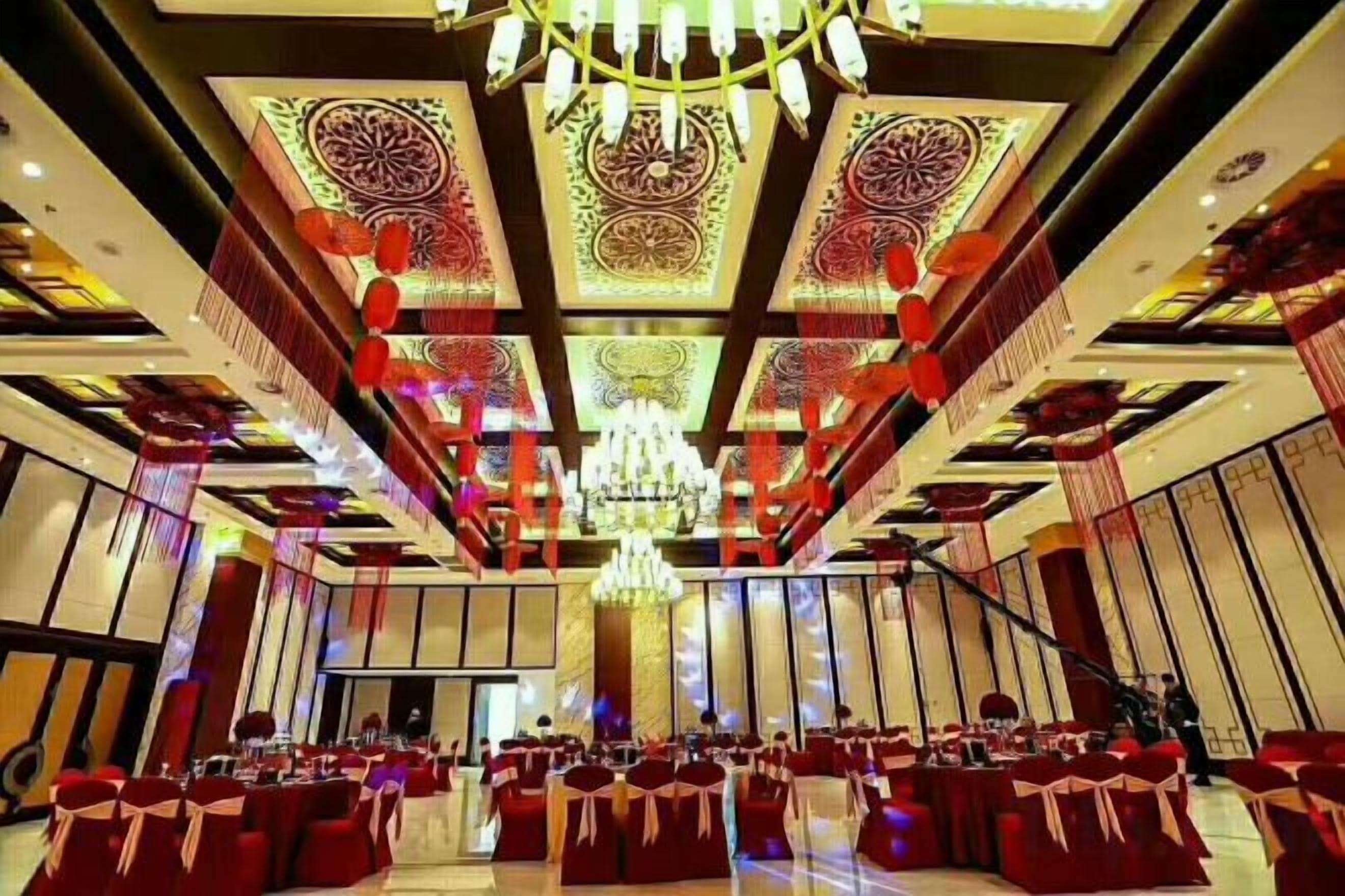 Un salón de banquetes con mesas y sillas dispuestas en filas, una araña y decoraciones geométricas en el techo.
