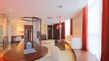 A modern hotel room with a round dining table, a white sofa, a small coffee table, and a large window with red curtains.
