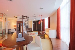 Una habitación de hotel moderna con una mesa redonda de comedor, un sofá blanco, una pequeña mesa de centro y un ventanal con cortinas rojas.