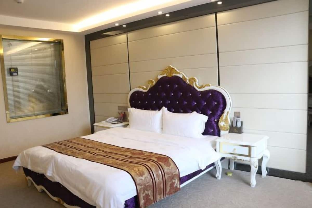 A hotel room with a large bed, a nightstand, a bedside table, and a window with blinds.