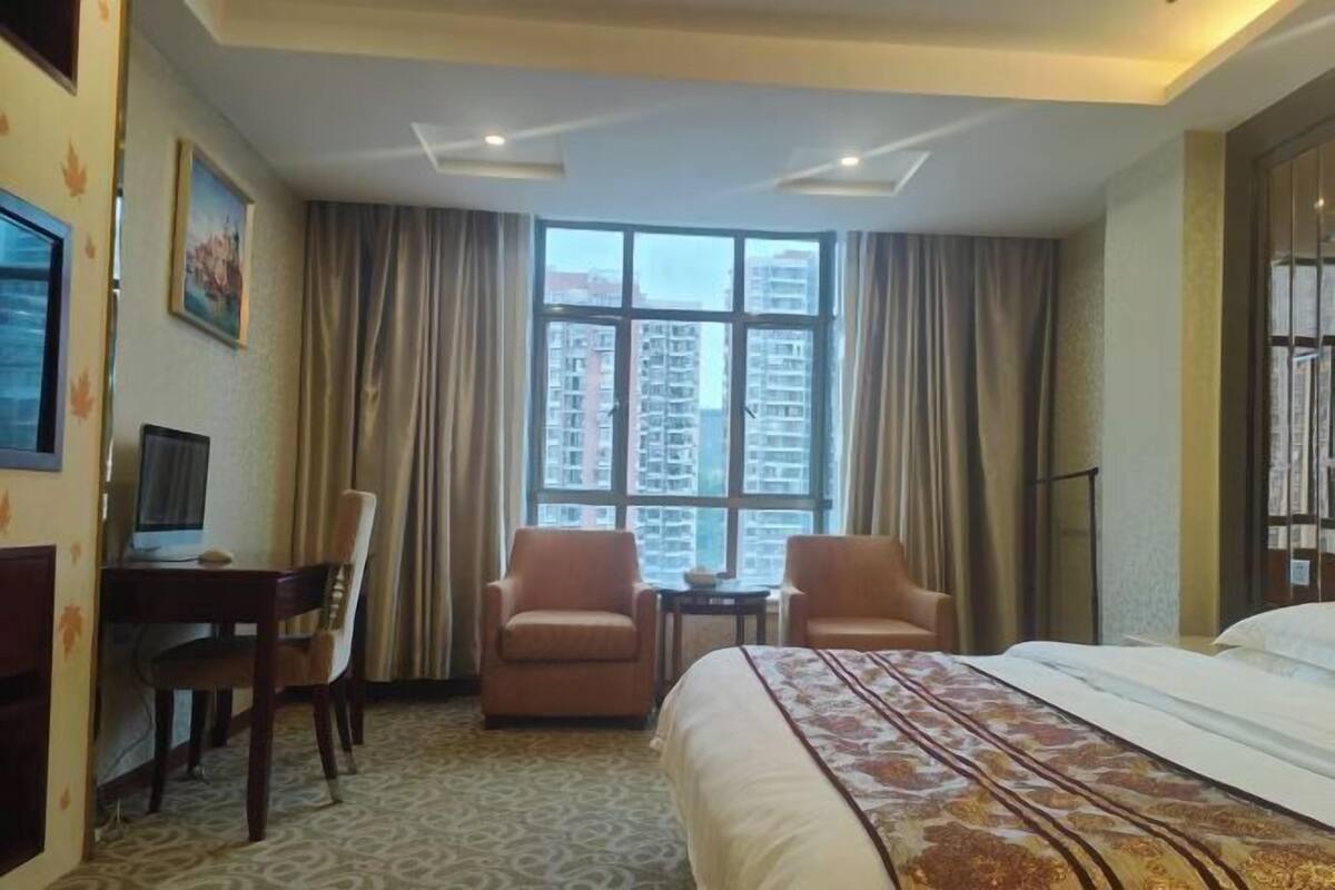 A hotel room with a bed, two armchairs, a desk with a computer, and a large window with a city view.