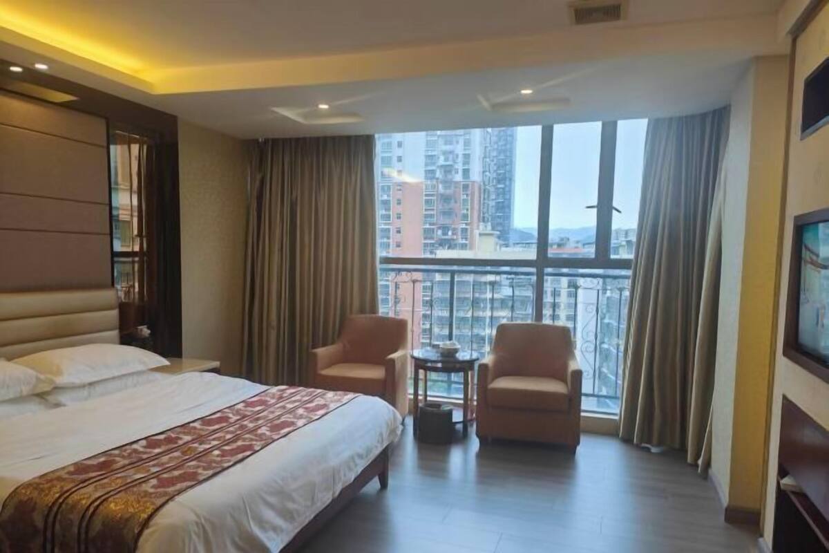 A hotel room with a large bed, two armchairs, a small table, and a large window with curtains.