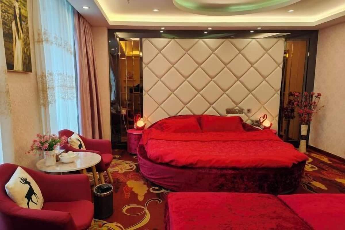 A bedroom with a large bed, red upholstered furniture, a round table with a vase of flowers, and a decorative wall panel.