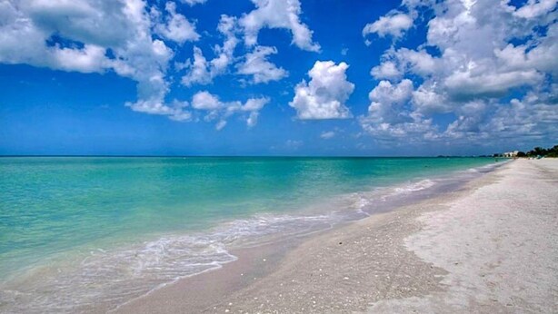 A beach with clear turquoise water, white sand, and a clear blue sky with scattered clouds.