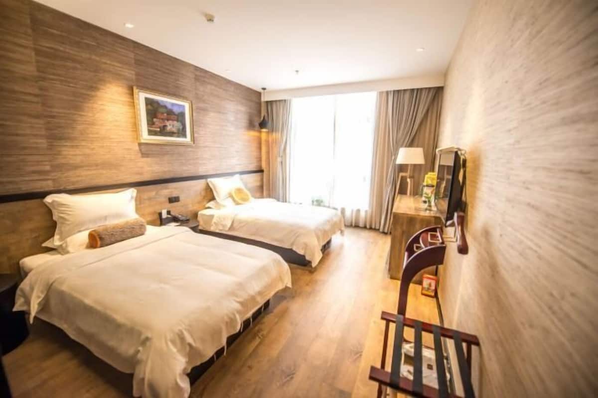 A hotel room with two beds, a wooden wall, a window with curtains, a nightstand, and a chair.