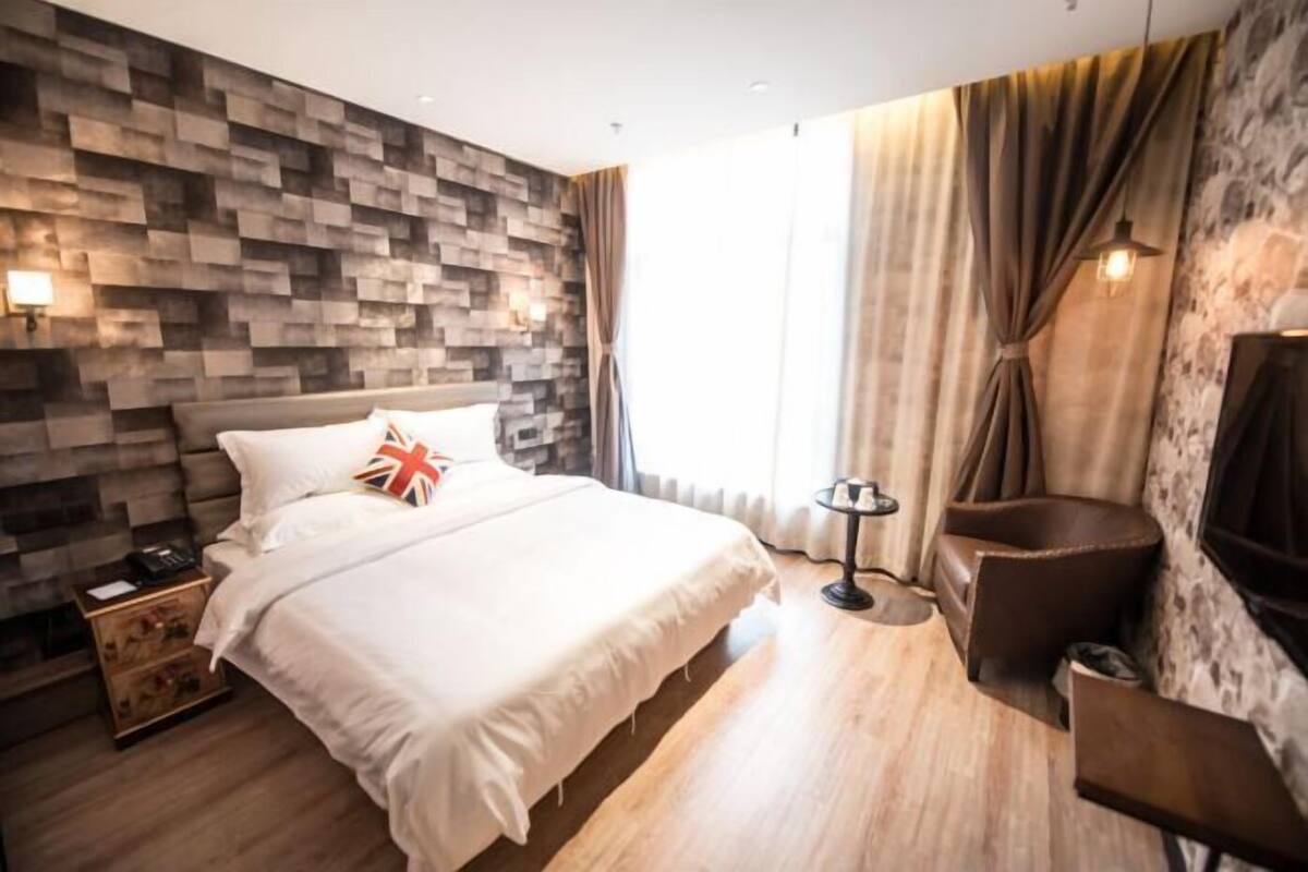 A modern hotel room with a wooden headboard, a bed with white linens, a bedside table with a lamp, a small round table, and a large window with curtains.